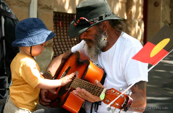 An Aboriginal musician kneels in front of a toddler allowing him to touch his guitar.