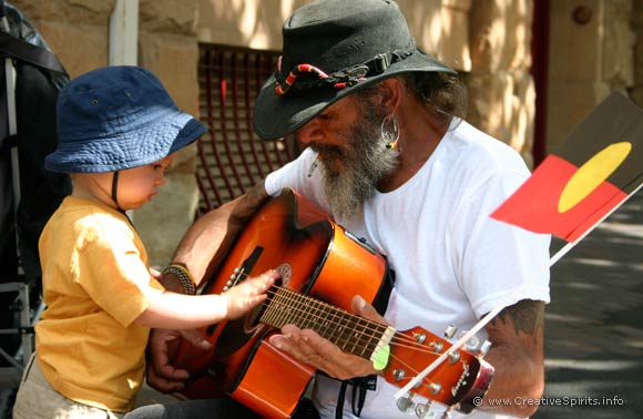 An Aboriginal man squatting on the footpath to let a toddler touch his guitar.