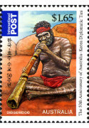 A didgeridoo player sits on red earth wearing a red head band and red short pants.