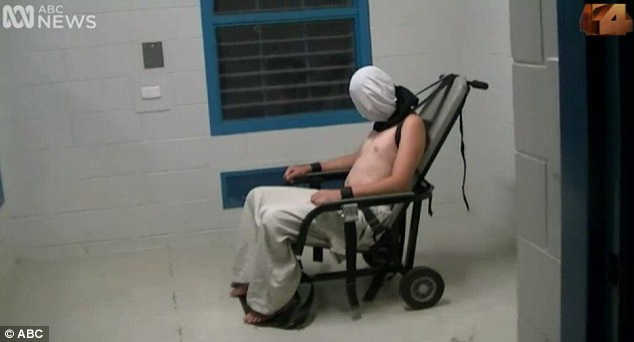 A bare-chested teenager sits bound and hooded in a chair.