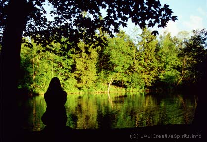 The dark outline of a woman sitting by herself at the shore of a lake.