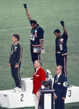 Three men stand on the victory dais, two black athletes raising their fist to the sky.