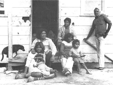 Essie's family gather in front of a simple wooden house entry.