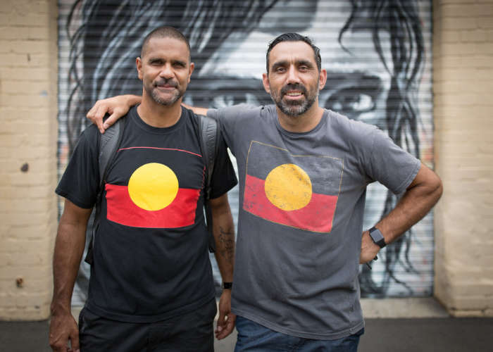 Michael O'Loughlin and Adam Goodes stand next to each other wearing t-shirts with an image of the Aboriginal flag.